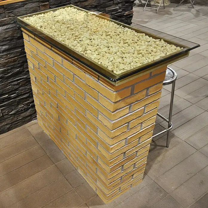 Esquina exterior Mediterráneo Barro - 24 x 12 x 5,5 cm - Un pedestal de ladrillos amarillos con una tapa de vidrio llena de piedras claras sirve como una elegante barra o mesa de exhibición.