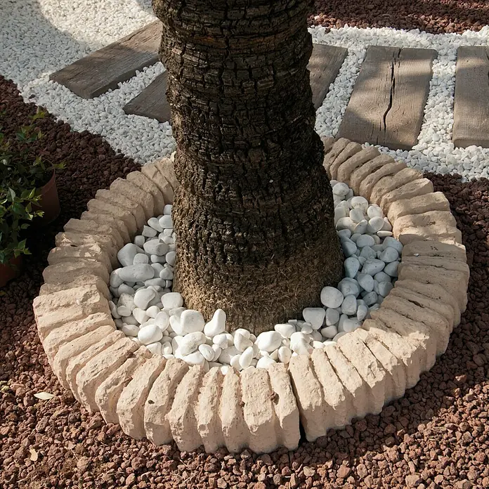 Paso para árboles Tostado - Piedra natural - Tronco de árbol con borde de piedra decorativa y guijarros blancos, rodeado de grava marrón.