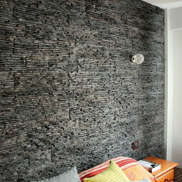 Revestimiento de piedra Galicia - Pizarra, Cantidad total: 4 pzs. - Primer plano de una pared decorativa con piedra apilada gris, una lámpara de pared con espejo y una mesita de noche con libros en el dormitorio.