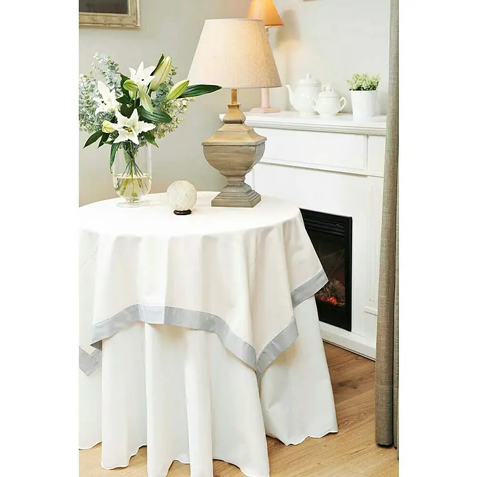 Mesa camilla redonda - 75 x 90 cm - Una mesa redonda con un mantel blanco de doble capa acentuado con gris, adornado con flores y una lámpara, destila elegancia.