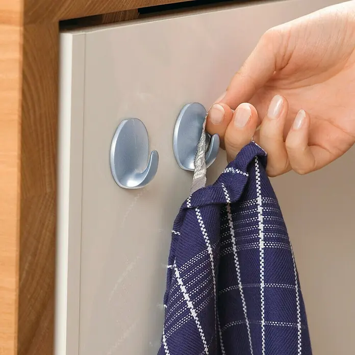 Tesa Powerstrips Gancho autoadhesivo Mini Redondo - Plateado - Primer plano de dos ganchos adhesivos plateados en la puerta de un armario, con una mano colgando un paño de cocina azul.