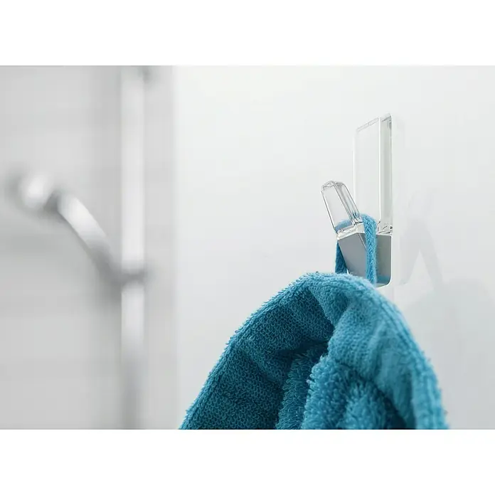 Tesa Powerstrips Gancho autoadhesivo - 2 ud., Transparente - Un toallero transparente y autoadhesivo con una toalla azul cuelga de una pared blanca en el baño.