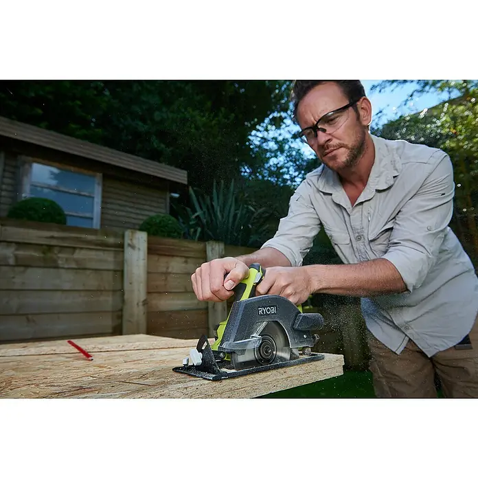 Ryobi ONE+ Maschinen-Set Combo Kit R18CK4G-252S - 18 V, 2 Akkus, 2 Ah - 5 Ah, 8 -tlg. - Ein Mann benutzt eine Ryobi Kreissäge, um ein Holzbrett zu schneiden, während er eine Schutzbrille trägt. Im Hintergrund ist ein Gartenhaus zu sehen.