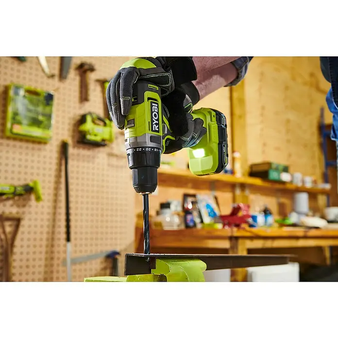 Ryobi ONE+ Maschinen-Set Combo Kit R18CK4G-252S - 18 V, 2 Akkus, 2 Ah - 5 Ah, 8 -tlg. - Ein RYOBI Akku-Bohrschrauber bohrt durch Metall, gehalten von behandschuhten Händen, mit Werkzeugen im Hintergrund.