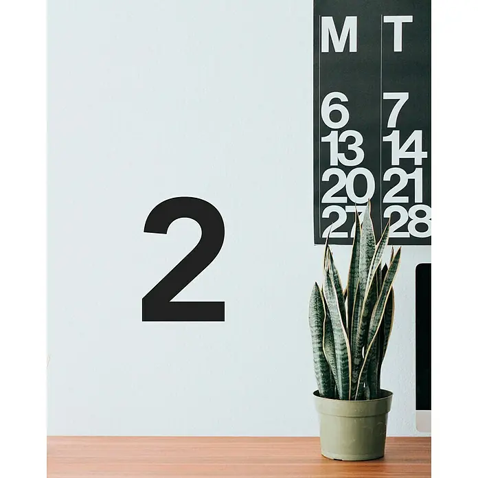 Vinilo de pared Número 2 - 4 x 9 cm, Negro, Plástico - Una planta de interior en maceta frente a una pared con un gran número '2' negro y un calendario.