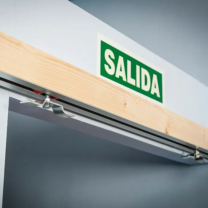 Carril de guía incrustado para puerta corredera - Largo: 2.000 mm, Capacidad de carga: 80 kg - Primer plano de una puerta corredera con un cartel de "Salida", que marca la puerta como salida. El foco está en la mecánica de la puerta.