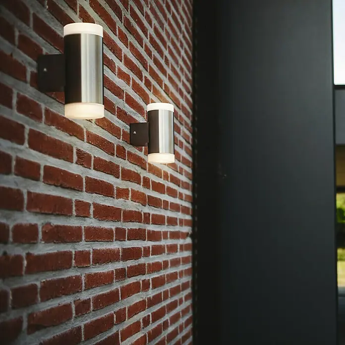 Lutec Aplique exterior LED Bilayer - 8 W, 13 x 13 x 11 cm, Plateado, IP44 - Imagen de dos lámparas de pared cilíndricas con carcasa de color plateado y luz blanca cálida en una pared de ladrillo.