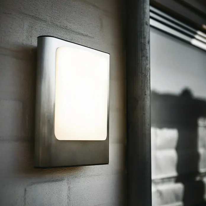 Lutec Aplique exterior LED Face - 16 W, 7 x 19 x 24 cm, Plateado, IP44 - Primer plano de una lámpara de pared rectangular con bordes redondeados que emite una luz cálida y difusa, montada en una pared.