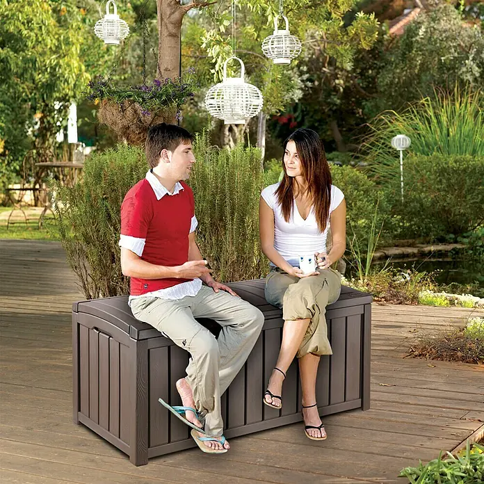 Keter Baúl para jardín Glenwood - Marrón, 65 x 128 x 61 cm, Polipropileno - Una pareja sentada en un banco de almacenamiento de jardín marrón hablando en el jardín. El banco ofrece un espacio práctico.