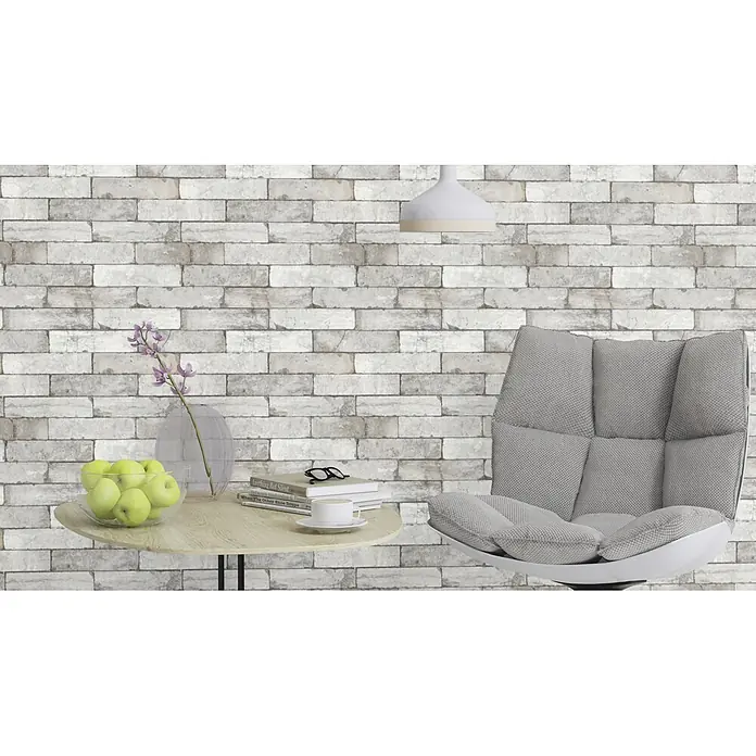 Rasch Vliestapete Factory 2 - Creme, Steinoptik, 10,05 x 0,53 m - Nahaufnahme eines modernen, grauen Sessels mit einem Beistelltisch mit Buch, Brille, Tasse, Blumenvase und einer Schale grüner Äpfel vor einer Ziegelwand.