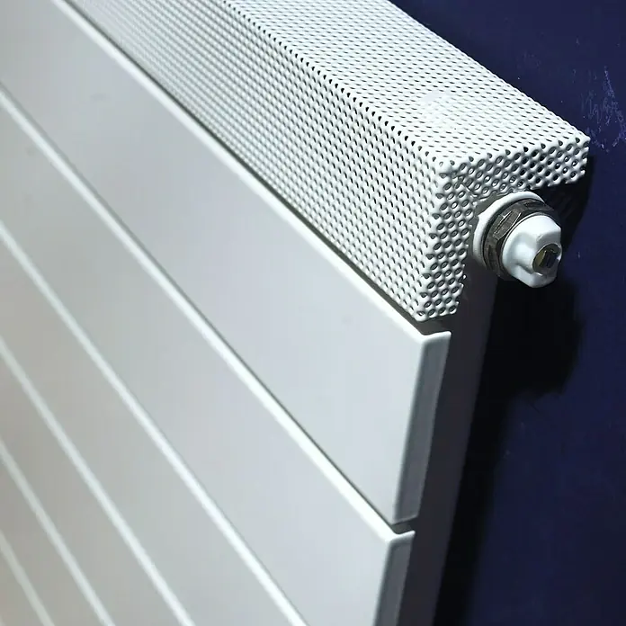 Ximax Designheizkörper Pep - B x H: 500 x 500 mm, Wärmeleistung bei Vorlauftemperatur von 75 °C (D: 50): 242 W - Nahaufnahme eines Heizkörpers mit weißer, perforierter Abdeckung und Thermostat vor einer blauen Wand.