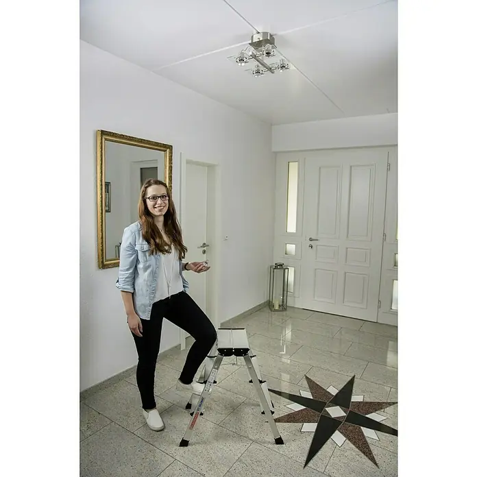Krause Monto Escalerilla plegable - Número de peldaños: 6 ud., Altura de trabajo: 2,65 m, Carga soportada: 150 kg, Aluminio - Una mujer está de pie en una escalera plegable Hailo en el pasillo, lista para cambiar la lámpara de techo. Práctica escalera para el hogar.