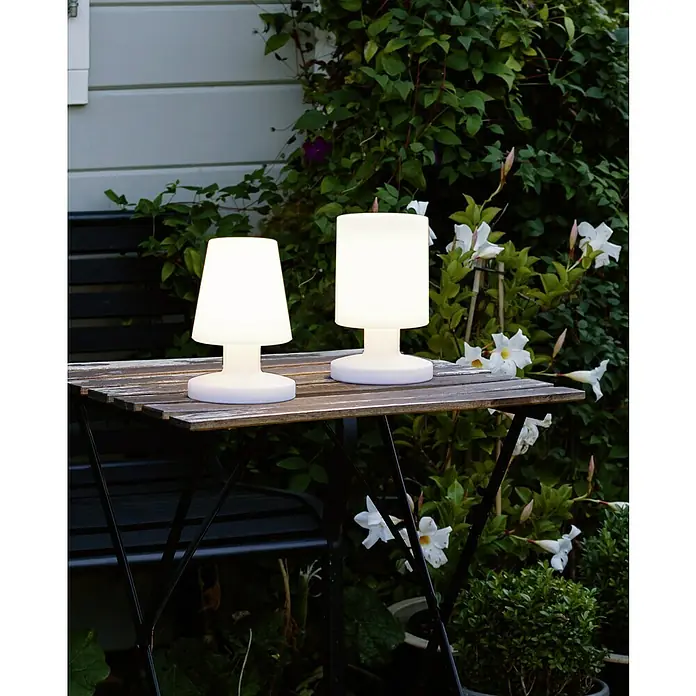 Reality Leuchten Akku-Tischleuchte Lora - 1,5 W, L x B x H: 13 x 13 x 20 cm, Weiß, Warmweiß - Zwei leuchtende Tischlampen stehen auf einem Holztisch im Freien und sorgen für stimmungsvolle Beleuchtung.