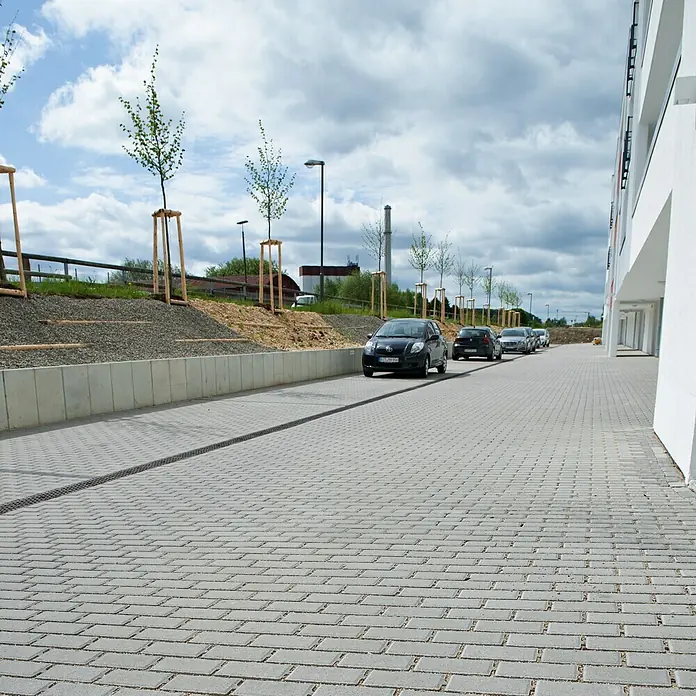 EHL Pflasterstein Drän - Grau, 20 x 10 x 8 cm, Rechteckig - Pflastersteine erstrecken sich entlang der Straße, wo Autos parken, mit jungen Bäumen und einem Wohnhaus im Hintergrund.