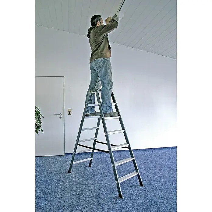 Krause Monto Doppelleiter SePro D - Anzahl Tritte: 14 Stk., Arbeitshöhe: 3,05 m, Aluminium - Eine Person steht auf einer grauen Leiter in einem Raum mit blauem Teppichboden und wechselt eine Glühbirne an der Decke.