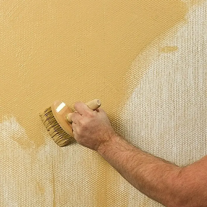 Claytec Lehm-Streichputz Clayfix - Kolumbagrau, 10 kg - Nahaufnahme einer Person, die mit einem breiten Pinsel eine grobe Leinwand mit gelber Farbe streicht, um die Textur hervorzuheben.