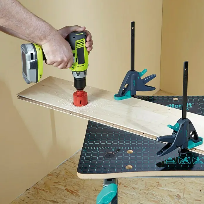 Wolfcraft Sargento 65-150-W - Anchura de sujeción: 120 mm - Un taladro inalámbrico con una sierra perforadora roja hace un agujero en una tabla de madera, sujeta por las abrazaderas Wolfcraft en un banco de trabajo.