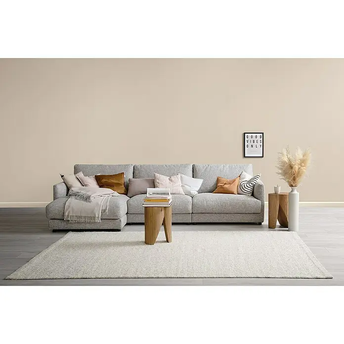 SCHÖNER WOHNEN-Farbe Wandfarbe Trendfarbe - Cosy, Matt, 5 l, Konservierungsmittelfrei - Ein graues Ecksofa mit Kissen und Decke auf einem hellen Teppich. Dekorative Elemente wie Beistelltische und ein Bild runden das Wohnzimmer ab.