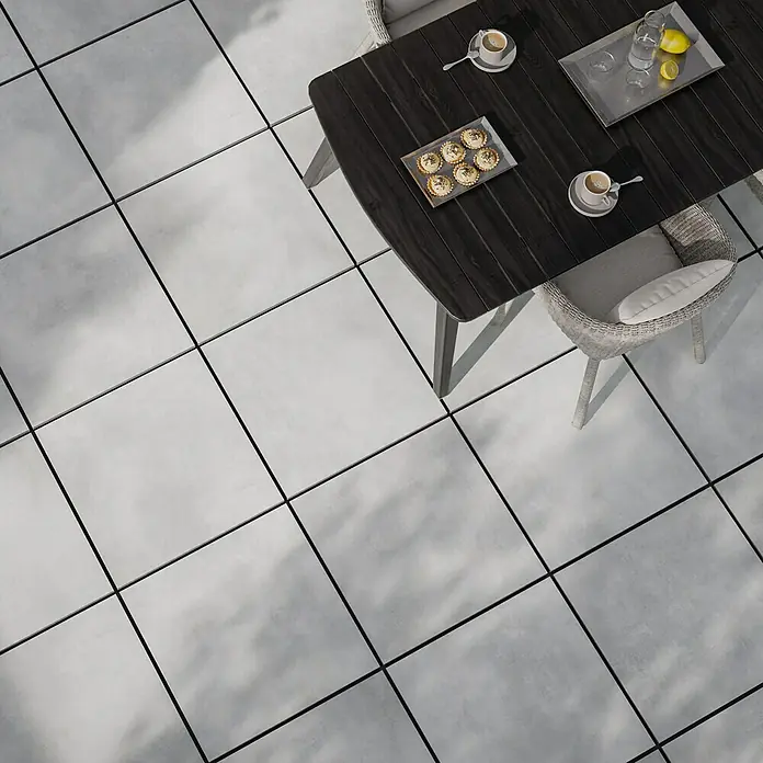 Terrassenfliese Dimension - 59,7 x 59,7 x 2 cm, Grau, Matt - Draufsicht auf einen dunklen Esstisch mit Geschirr und Speisen auf hellgrauen Fliesen, umgeben von einem Rattansessel mit Kissen.
