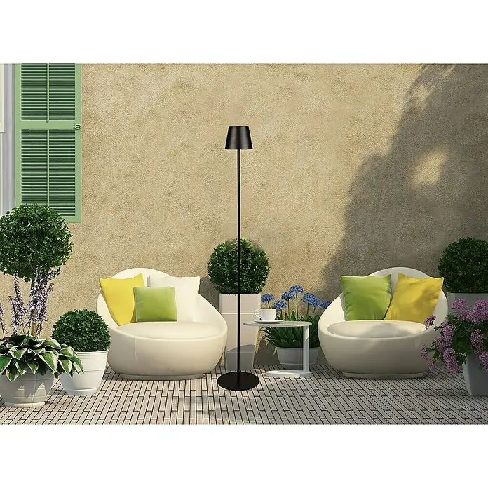Starlux Paola Akku-Stehleuchte - Schwarz, RGBW, 3 W, IP44 - Stimmungsvolle Außenansicht mit einer eleganten schwarzen Stehlampe, umgeben von stilvollen Möbeln und üppigem Grün.