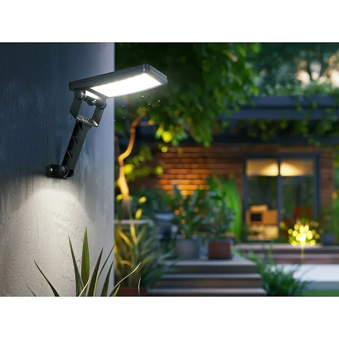 Garza Aplique solar para exterior Carvalho - L x An x Al: 33 x 11 x 4,3 cm, Negro, 500 lm - Una luz solar está montada en una pared, iluminando un jardín con plantas, una casa y escalones.