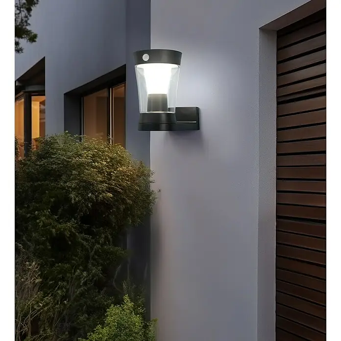Garza Hércules Aplique solar para exterior - L x An x Al: 12,2 x 22,3 x 19,4 cm, Negro, IP54, 450 lm - Una moderna lámpara de pared negra con sensor de movimiento ilumina la pared de una casa, brindando seguridad y estilo al exterior.