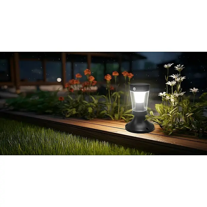 Garza Hércules Sobremuro solar LED - L x An x Al: 18 x 18 x 26,9 cm, Negro, IP54, 450 lm - Una luz solar en un patio de madera junto a flores y hierba, creando un ambiente acogedor en el jardín.