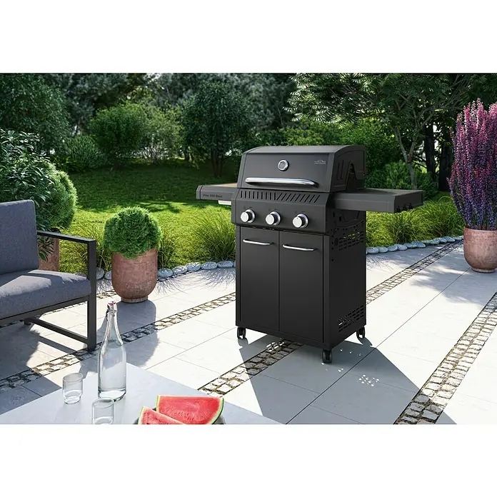 Kingstone Gasgrill Diner 3000 Beast - Grillfläche (B x T): 63,7 x 43 cm, 10,5 kW, Anzahl Brenner: 3 Stk. - Eleganter, schwarzer Napoleon Grill auf einer Terrasse mit Steinfliesen, umgeben von üppigem Grün. Ideal für sommerliche Grillabende.