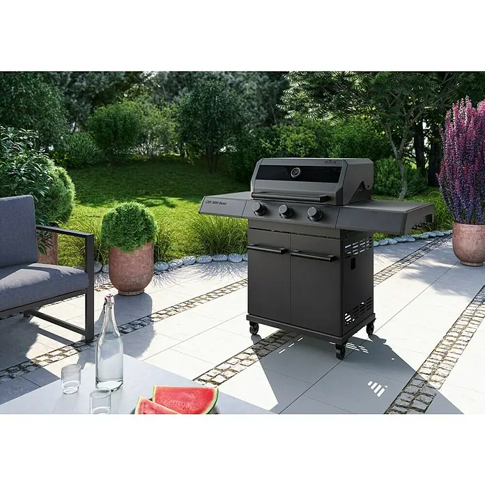 Kingstone KMS Gasgrill Cliff 3000 Beast Black Edition - Grillfläche (B x T): 61 x 45 cm, 10,5 kW, Anzahl Brenner: 3 Stk. - Draußen steht ein moderner schwarzer Grill der Marke Cliff 2000 Beast auf einer Terrasse, umgeben von üppigem Grün.