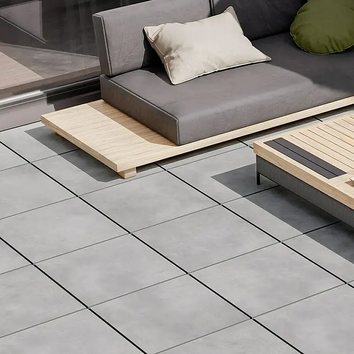 Terrassenfliese Dimension - 59,7 x 59,7 x 2 cm, Grafit, Matt - Nahaufnahme eines grauen Sofas mit hellen Kissen auf einer Terrasse mit grauen Fliesen.