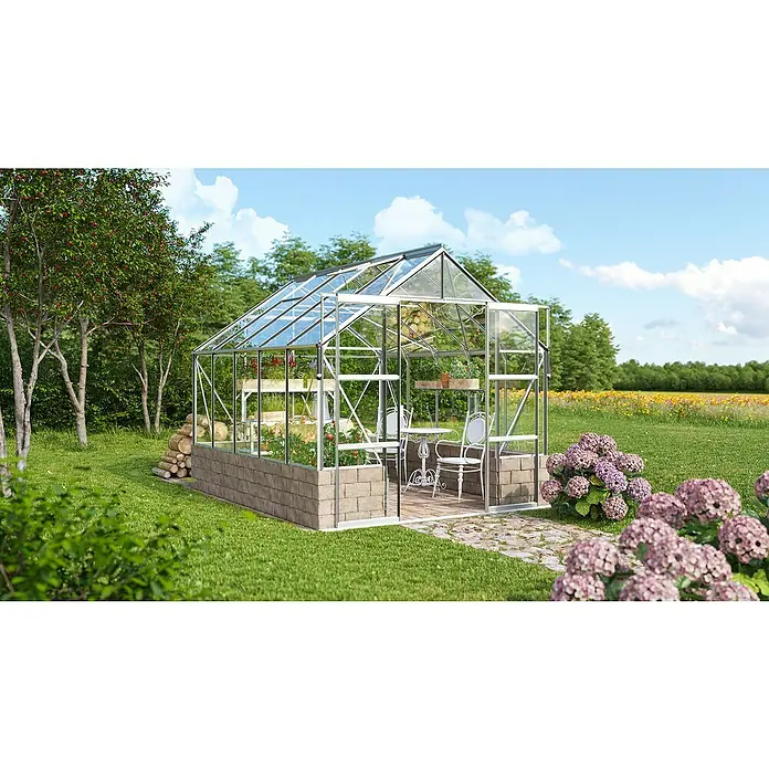 Vitavia Gewächshaus Cassandra 8300 - Außenmaß inkl. Dachüberstand (B x T): 257 x 323 cm, 3 mm, Aluminium - Ein schönes Gewächshaus mit Glaseinsätzen im Garten, umgeben von Grün und Blumen. Es verfügt über ein Beet, einen Tisch und Stühle.