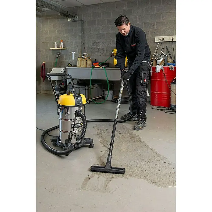 PowerPlus Aspirador en seco y húmedo eléctrico POWX3240 - 1.200 W, Volumen de recipiente: 30 l, Con bolsa - Un hombre limpia el piso de un garaje con una aspiradora para sólidos y líquidos Powerplus amarilla y plateada.