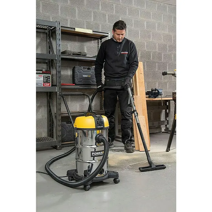 PowerPlus Aspirador en seco y húmedo eléctrico POWX3240 - 1.200 W, Volumen de recipiente: 30 l, Con bolsa - Un hombre usa una aspiradora Powerplus con tapa amarilla y carcasa cromada en un taller para aspirar aserrín.