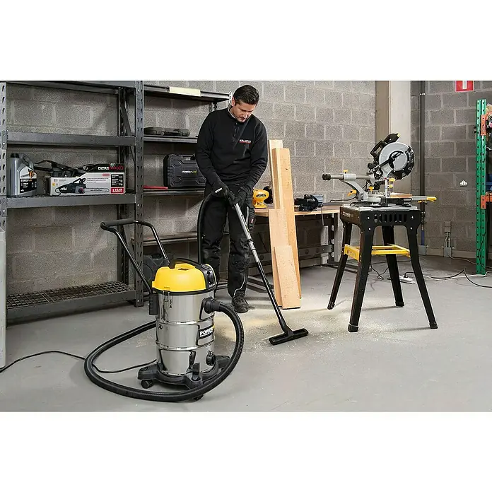 PowerPlus Aspirador en seco y húmedo eléctrico POWX3240 - 1.200 W, Volumen de recipiente: 30 l, Con bolsa - Un hombre utiliza una aspiradora Powerplus para limpiar un taller, con estanterías, una sierra y madera.