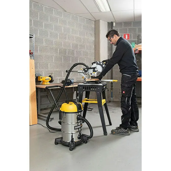 PowerPlus Aspirador en seco y húmedo eléctrico POWX3240 - 1.200 W, Volumen de recipiente: 30 l, Con bolsa - Un hombre usa una sierra ingletadora Powerplus sobre un banco de trabajo, con una aspiradora cerca para la extracción de polvo.