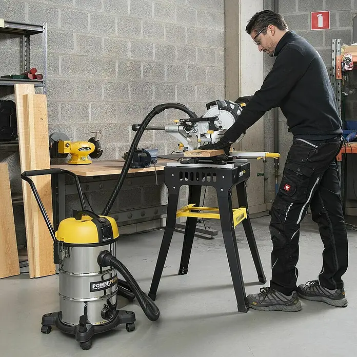PowerPlus Aspirador en seco y húmedo eléctrico POWX3240 - 1.200 W, Volumen de recipiente: 30 l, Con bolsa - Un hombre opera una sierra de inglete mientras una aspiradora de taller amarilla y plateada se encuentra cerca, manteniendo el espacio limpio.