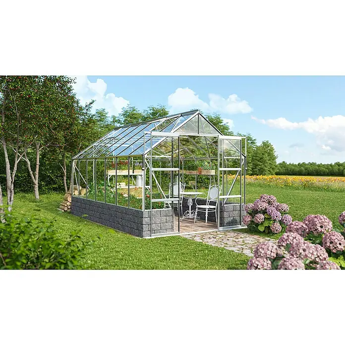Vitavia Gewächshaus Cassandra 11500 - Außenmaß inkl. Dachüberstand (B x T): 257 x 448 cm, 3 mm, Aluminium - Die Aufnahme zeigt ein Gewächshaus im Garten, umgeben von Blumen und Bäumen, mit dem Fokus auf die Architektur des Gewächshauses.