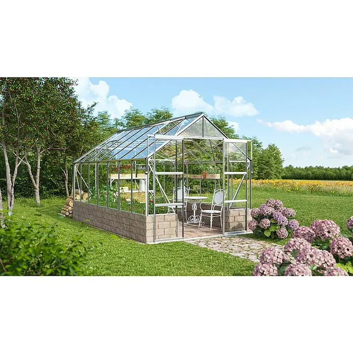 Vitavia Gewächshaus Cassandra 11500 - Außenmaß inkl. Dachüberstand (B x T): 257 x 448 cm, 3 mm, Aluminium - Das Bild zeigt ein Gewächshaus mit Pflanzen und Gartenmöbeln, umgeben von Blumen und Bäumen, unter einem hellblauen Himmel.