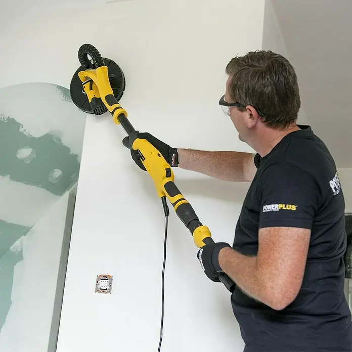 PowerPlus Lijadora de pared y techo POWX04761 - 710 W, Diámetro disco de lija: 225 mm - Un hombre lija una pared con una lijadora de paneles de yeso POWERPLUS amarilla. Este dispositivo es telescópico para lijar paredes fácilmente.