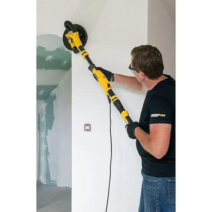 PowerPlus Lijadora de pared y techo POWX04761 - 710 W, Diámetro disco de lija: 225 mm - Un hombre lija una pared con una lijadora de paneles de yeso amarilla con mango telescópico, lo que permite un trabajo eficiente en grandes superficies.