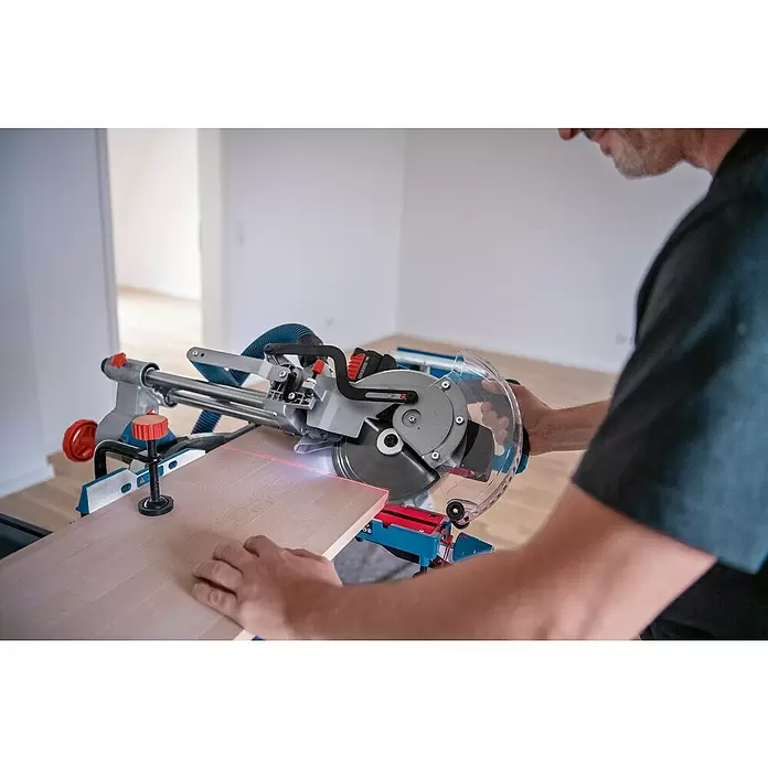 Bosch Professional AMPShare 18V Akku-Kapp- & Gehrungssäge GCM 18V-216 D - 18 V, Ohne Akku, 5.000 U/min - Mann schneidet mit einer grau-blauen Gehrungssäge und rotem Laser eine Holzplanke. Die Säge hat einen transparenten Blattschutz.