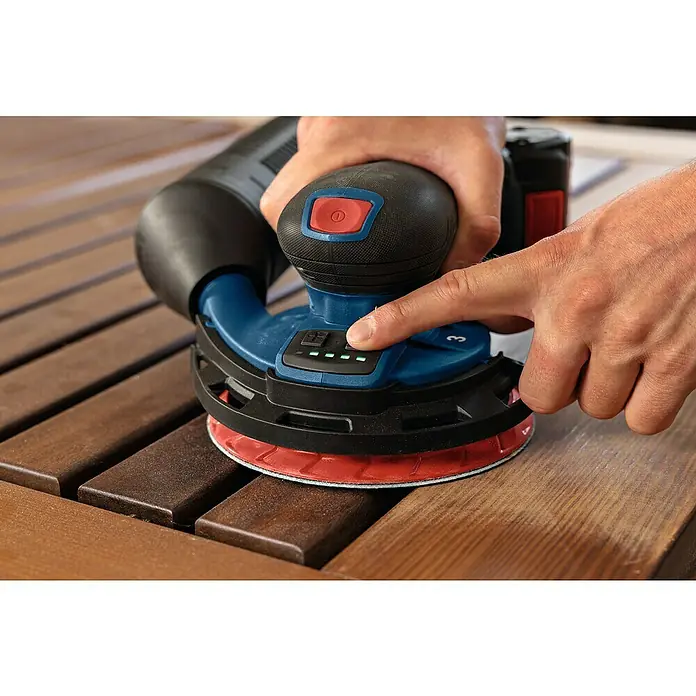 Bosch Professional AMPShare 18V Akku-Exzenterschleifer GEX 18V-150-3 - 18 V, Ohne Akku, Durchmesser Schleifteller: 150 mm - Nahaufnahme eines Akku-Exzenterschleifers beim Schleifen einer Holzoberfläche, wobei der Finger einen Knopf drückt, um die Geschwindigkeit einzustellen.