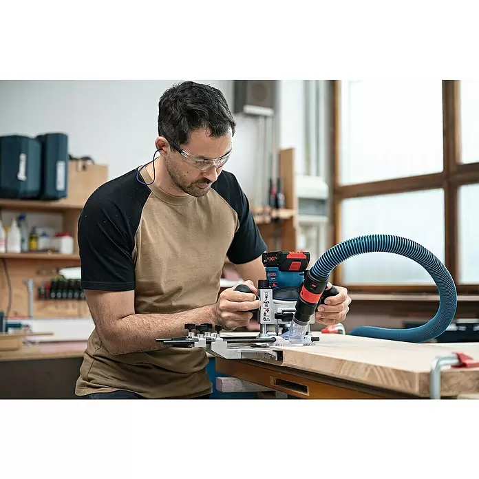 Bosch Professional AMPShare 18V Akku-Kantenfräser GKF 18V-8 - 18 V, Anzahl Akkus: Ohne Akku, Leerlaufdrehzahl: 10.000 U/min - 30.000 U/min - Ein Handwerker nutzt eine Bosch Akku-Oberfräse und einen blauen Staubabsaugschlauch für präzise Holzbearbeitung.