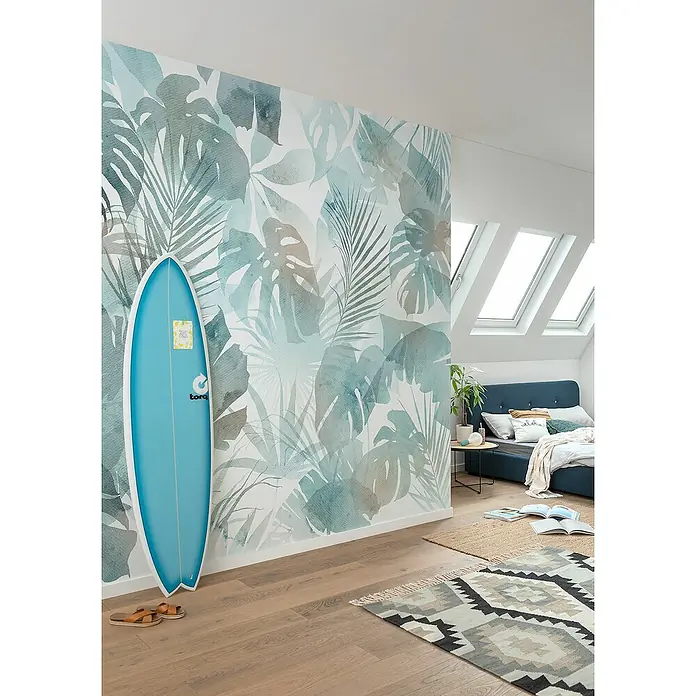 Komar Flair Fototapete Aqua Flora - B x H: 300 x 250 cm, Vlies - Ein hellblaues Surfbrett lehnt an einer Wand mit tropischem Blätterdruck in einem sonnigen Schlafzimmer mit Dachfenstern.