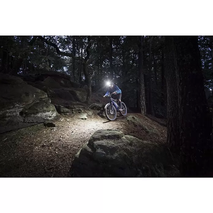 Fischer LED-Leuchte Akku - Beleuchtungsstärke: 1.600 lx - Helle Stirnlampe beleuchtet den Weg für einen Radfahrer beim Mountainbiken durch einen dunklen, felsigen Wald bei Nacht.
