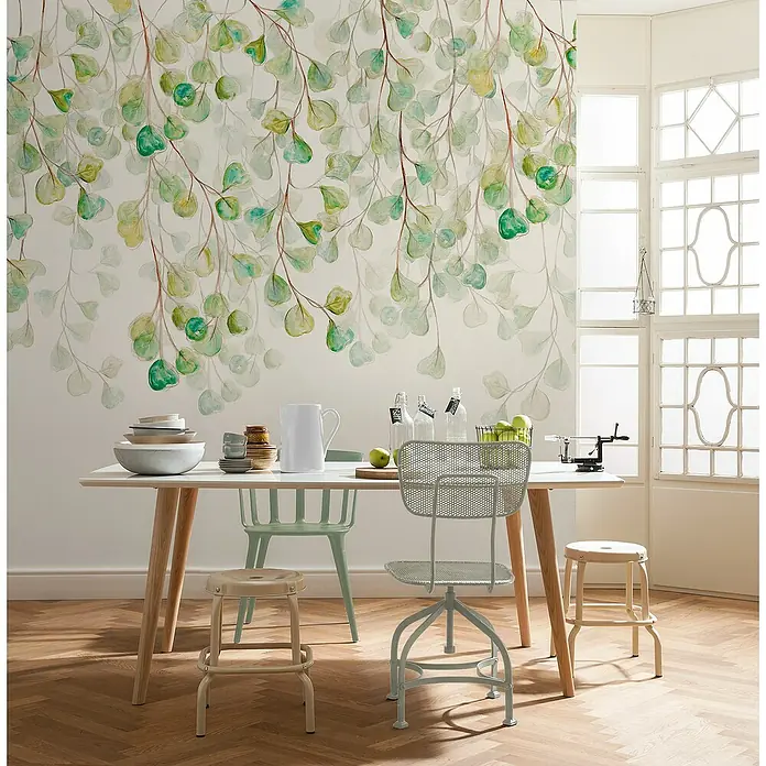 Komar Flair Fototapete Douche de Feuilles - B x H: 300 x 250 cm, Vlies - Ein Esstisch mit Geschirr, Stühlen und Hockern vor einer Wand mit Blattmuster-Tapete.