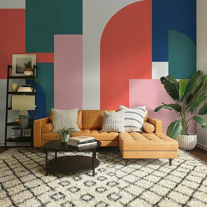 Komar Flair Fototapete Geometric Colours - B x H: 350 x 250 cm, Vlies - Wohnzimmer mit ockerfarbenem Ledersofa, Couchtisch und Bücherregal vor einer bunten geometrischen Wand.