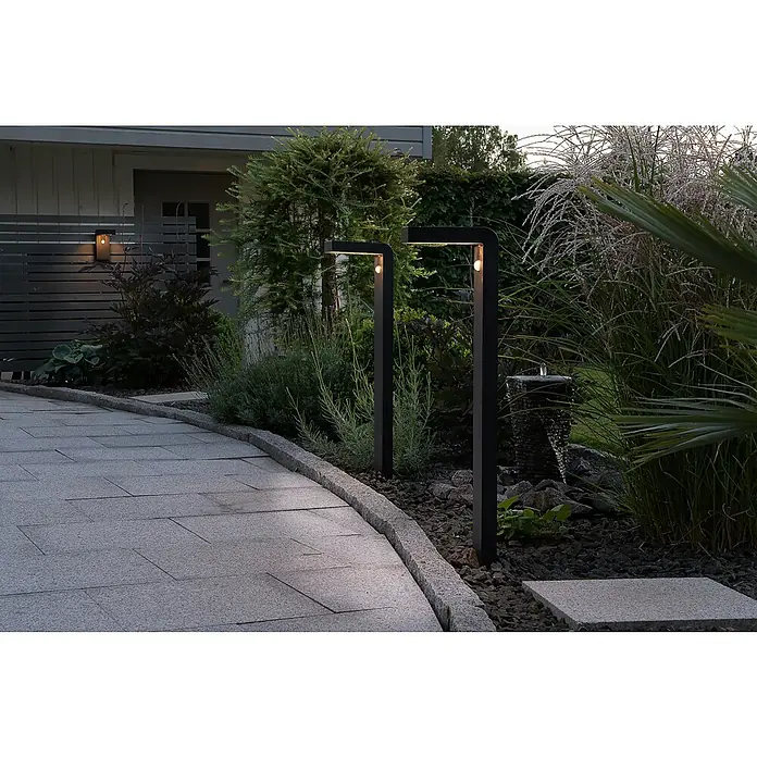 Eglo Aplique solar para exterior Baracconi - 18 W, Negro, IP44, 300 lm - Dos modernas luces de jardín de metal negro iluminan un camino pavimentado, rodeado de exuberante vegetación y una cascada decorativa.