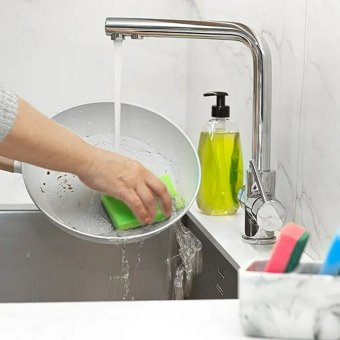 Vileda Glitzi Topfreiniger Colors 2+1 - 3 Stk. - Frau reinigt eine Bratpfanne mit einem grünen Schwamm unter einem modernen Wasserhahn. Spülmittel und Spülutensilien stehen bereit.
