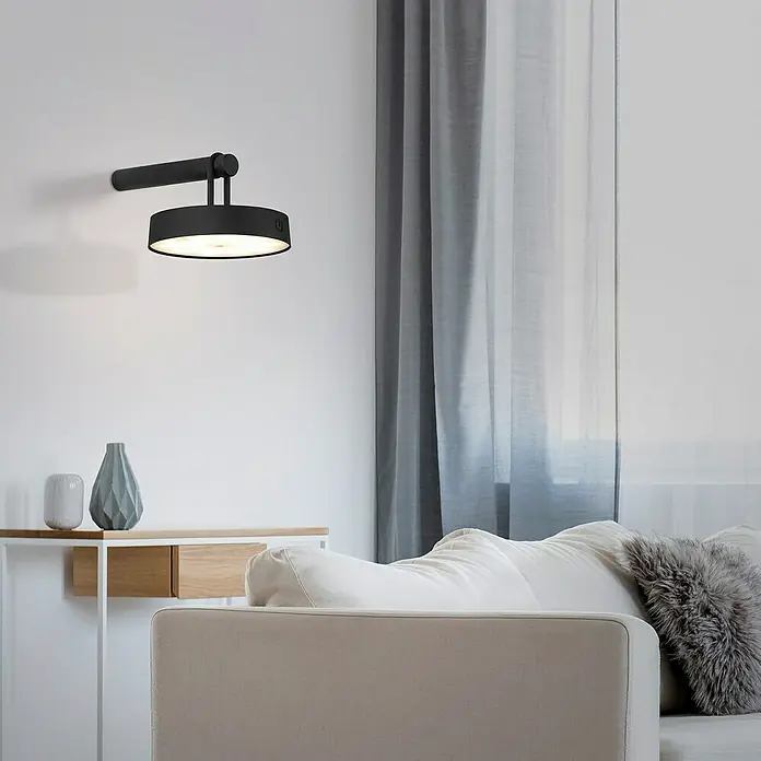 Just Light Aplique de pared con batería Arma - 12,8 x 19 x 8,7 cm, Negro, Blanco cálido, 15 W, IP44 - Una lámpara de pared negra moderna con una pantalla redonda emite una luz cálida en una sala de estar elegante con jarrón y sofá.